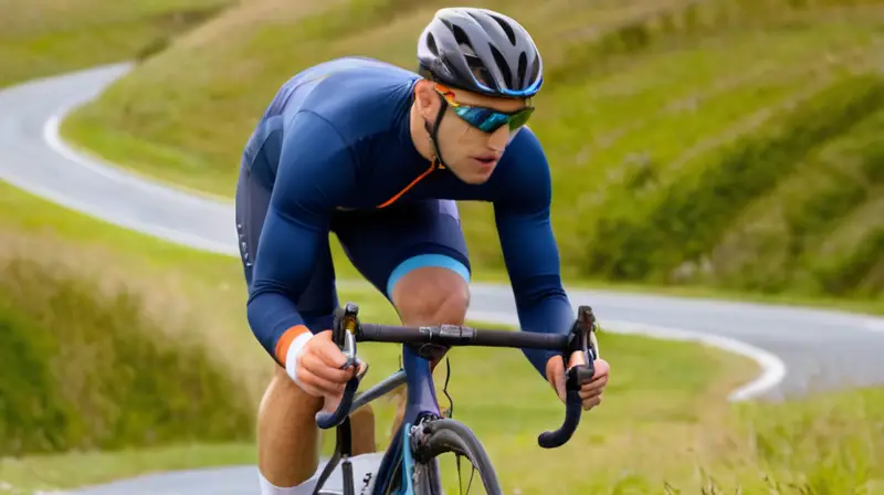 Jersey de ciclismo ajustado en azul marino con detalles naranjas, paneles de malla, y un ciclista en movimiento sobre un fondo difuso