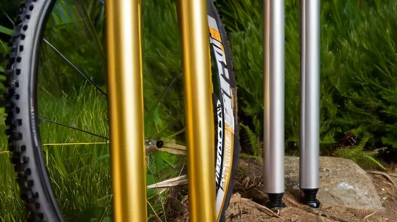 Dos horquillas de suspensión de bicicleta de montaña, una dorada y otra plateada, destacan sobre una superficie de madera rústica